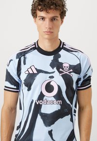 Svetlomodrý športový dres s čiernymi šmuhami, čiernymi akcentmi a čiernym golierom. Obsahuje logá: ružová lebka s kosťami.