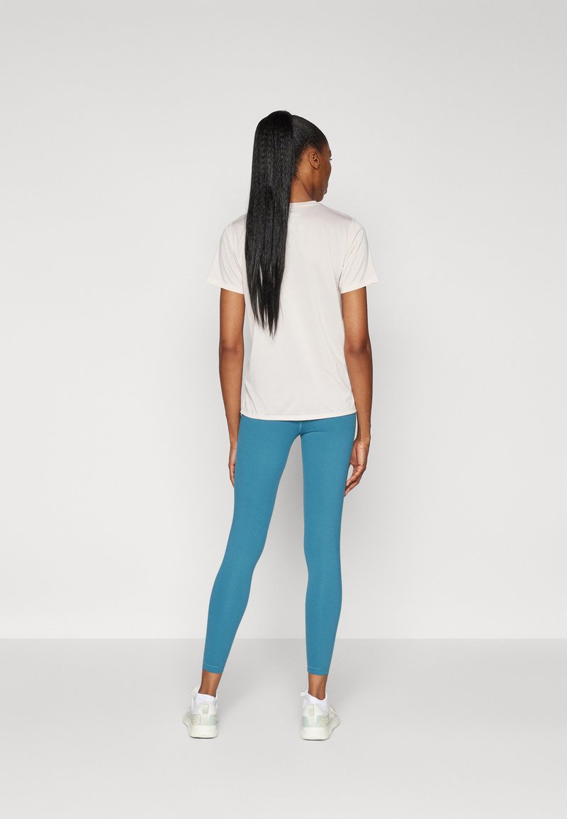 Une personne portant une chemise à manches courtes rose clair et des leggings bleus, avec des cheveux noirs raides attachés en arrière, vue de dos.