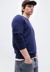 Νεαρός άνδρας με καφέ καπέλο, μπλε σκούρο φούτερ πάνω από λευκό πουκάμισο και ανοιχτόχρωμο μπλε τζιν, στέκεται με τα χέρια στις τσέπες του μπροστά σε λευκό φόντο.