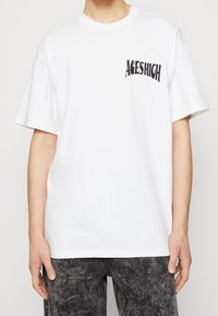 Biała bawełniana koszulka z czarnym, zakrzywionym nadrukiem "ACESHIGH" na lewej piersi. Krótkie rękawy, okrągły dekolt, luźny krój.