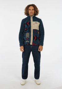 Mann mit lockigem Haar, der steht und eine blaue Fleecejacke mit Muster, dunkle Jeans und weiße Turnschuhe trägt, vor einem schlichten weißen Hintergrund.