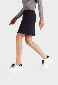 Sneakers platform bianche con suola texturizzata, dettaglio del tallone nero e lacci neri. Il modello indossa un maglione grigio e una gonna blu navy, in movimento.