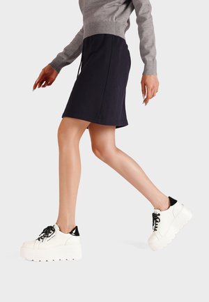 Sneakers platform bianche con suola texturizzata, dettaglio del tallone nero e lacci neri. Il modello indossa un maglione grigio e una gonna blu navy, in movimento.