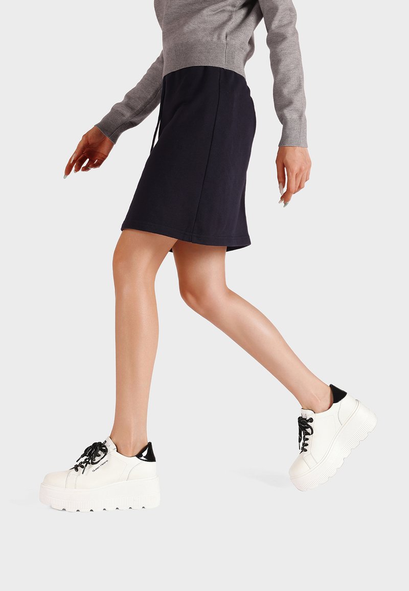 Sneakers platform bianche con suola texturizzata, dettaglio del tallone nero e lacci neri. Il modello indossa un maglione grigio e una gonna blu navy, in movimento.