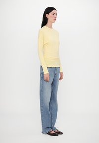 Langärmliges, hellgelbes Oberteil aus weichem Stoff, kombiniert mit locker sitzenden, hellblauen Jeans. Model trägt flache, dunkle Schuhe.