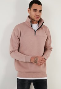 Sudadera rosa claro con un alto cuello, cierre de media cremallera, puños y dobladillo acanalados. Fabricada con tela suave, llevada sobre una camiseta blanca.
