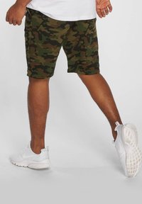 Pantaloni corti con stampa camouflage in verde, marrone e nero. Design aderente con tasche laterali e cuciture a vista. Indossati con scarpe da ginnastica bianche.