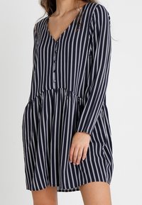 Granatowa sukienka z pionowymi białymi paskami, dekoltem w serek, długimi rękawami, detalami w postaci guzików oraz rozkloszowaną częścią spódnicy z marszczoną talią.