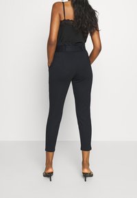 Haut sans manches noir avec un bord en dentelle, associé à un pantalon ajusté bleu marine doté d'une taille ceinturée et de poches latérales. Texture lisse.