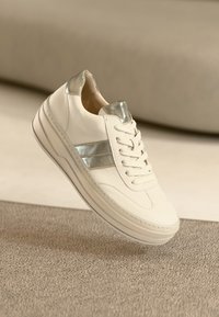 White leather sneaker with metallic silver accents, round toe, lace-up closure, and textured rubber sole. Minimalist design with smooth finish.