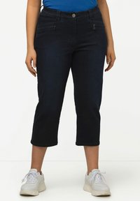Jeans capri azul oscuro con una textura suave, con una cintura de media altura y dos bolsillos frontales con cremallera. Combinados con zapatillas deportivas blancas.