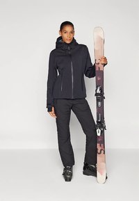 Musta vedenpitävä hiihtotakki vetoketjulla edessä, sopivat housut ja mustat hiihtomonot. Pitelee ruskeita ja vaaleanpunaisia suksia siteineen.