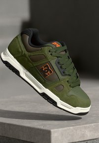 Green suede and textile sneaker with black accents and orange logo, featuring a cushioned sole and textured surface details.