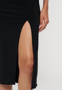 La main d'une femme reposant sur la cuisse, vêtue d'une jupe noire avec une fente haute montrant le haut de la jambe sur un fond uni.