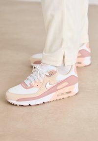 Baltos, rožinės ir smėlinės spalvos Nike Air Max sportbačiai su tinkliniu ir odiniu viršumi, apvaliu nosimi ir matoma oro pagalvėle pado viduje.