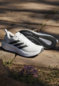 Chaussures de course blanches avec des bandes noires et des semelles rembourrées reposant sur un pavé en pierre à l'extérieur avec de petites plantes à proximité.