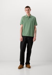 Žalias polo marškinėliai su balta apykaklės apdaila, derinami su tamsiais džinsais ir rudais slip-on batais. Polo ant krūtinės turi logotipą.
