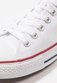 Gros plan d'une basket en toile blanche avec lacets blancs, œillets argentés, bande rouge au-dessus de la semelle en caoutchouc et bout renforcé blanc texturé.