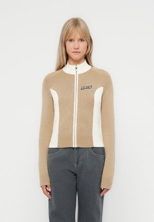 Beige en witte zip-up trui met hoge kraag, geribbelde textuur en logo op de borst. Heeft een getailleerd ontwerp en lange mouwen.