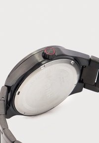 Πίσω μέρος μαύρου ρολογιού καρπού Hugo από ανοξείδωτο ατσάλι που δείχνει το κορόνα με κόκκινο ακцент και χαραγμένες λεπτομέρειες στη θήκη, με γωνία σε λευκό φόντο.