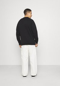 Felpa nera con maniche raglan, polsini e orlo a costine, indossata sopra pantaloni cargo bianchi, vista da dietro. Texture del tessuto liscia.