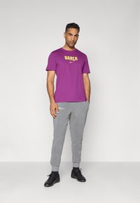 Vijolična majica s "BARÇA" in logotipom Nike v rumeni barvi, v kompletu s sivo prilegajočimi trenirkami in črnimi športnimi čevlji, prikazana na nevtralni podlagi.