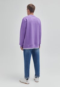 Jeune homme portant un sweat-shirt lavande, un jean bleu et des baskets blanches, debout de dos devant un fond clair uni.