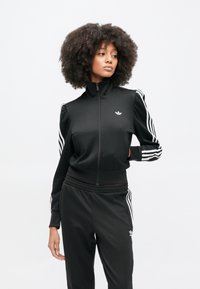 Μαύρη αθλητική φόρμα με μπουφάν με φερμουάρ, ψηλό γιακά και λευκές ρίγες στα μανίκια. Διαθέτει μικρό λογότυπο στο στήθος και ελαστική μέση.