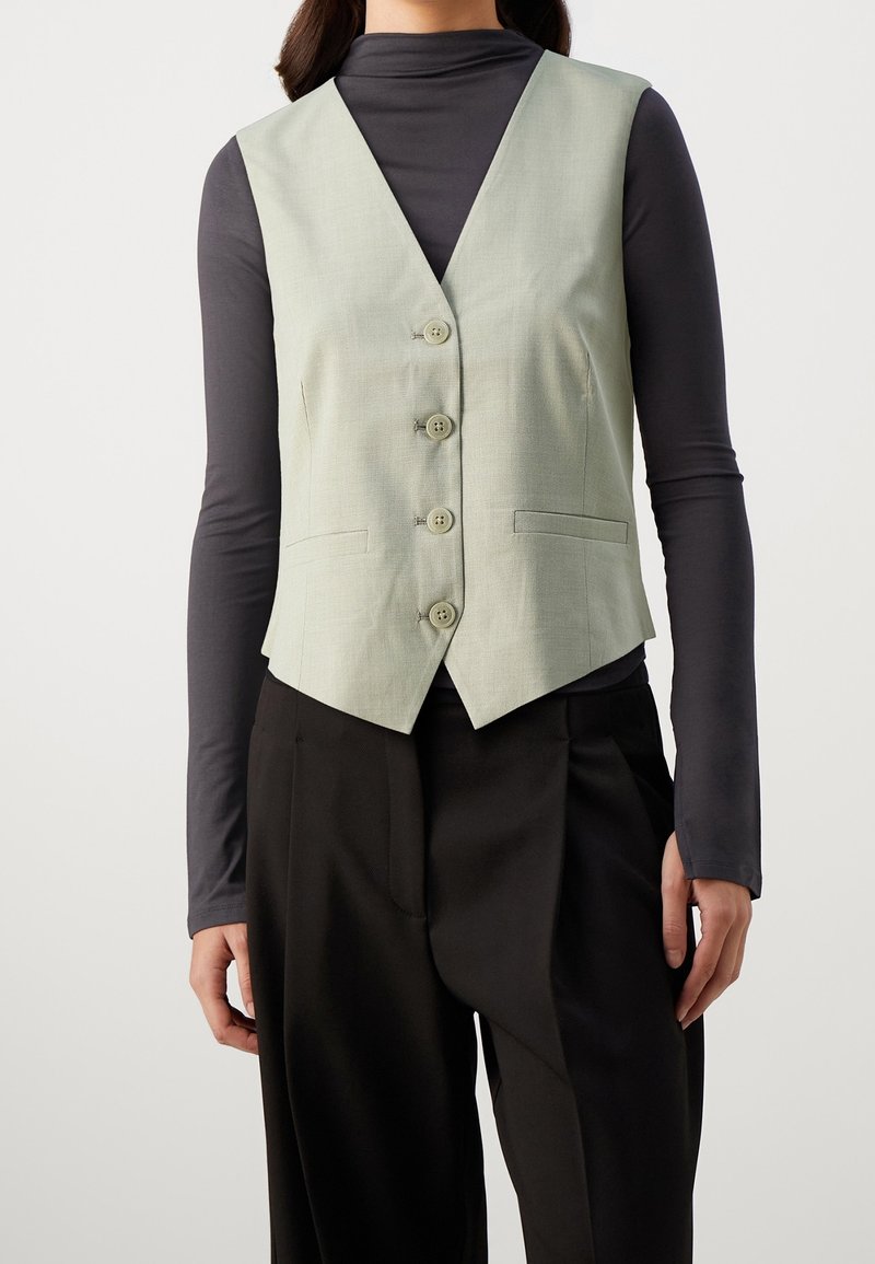 Hellgrüner, taillierter Weste mit V-Ausschnitt, sechs Knöpfen und zwei Seitentaschen, getragen über einem schwarzen Langarmoberteil.