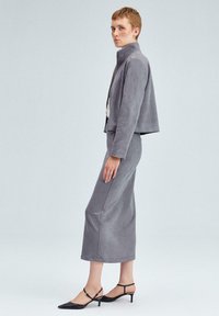 Hallast faux-suede jakk, millel on tõusnud krae, ühesuguse midi seeliku ja mustade teravatipuliste kontsakingadega, millel on pahkluu rihmad.