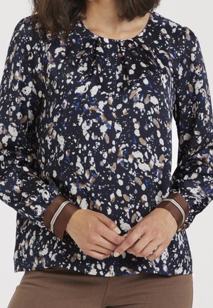 Femme portant un chemisier bleu marine avec des taches abstraites blanches, brunes et bleues, et des poignets bruns, les mains jointes devant.