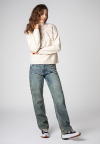 CremefarbenerStrickpullover mit hohem Kragen und dekorativem Muster, kombiniert mit lässigen hellblauen Jeans und schwarzen Sneakers.