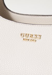 Détail d'un sac en cuir blanc texturé avec le lettrage doré du logo « GUESS EST 1981 » sur le bord supérieur lisse.