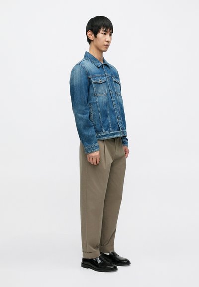 Veste en denim avec deux poches poitrine, lavage bleu clair, matériau robuste. Associée à un pantalon beige ample et des chaussures en cuir noir.