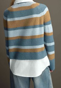 Pull rayé en laine bleu, bleu clair et marron, avec un ourlet et des poignets côtelés. Porté par-dessus une chemise blanche à col, associé à un jean en denim clair.