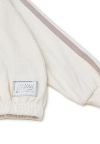 Sudadera de felpa blanca con una etiqueta de logo plateado, con puños acanalados y una franja beige a lo largo de la manga. Textura suave y diseño casual.