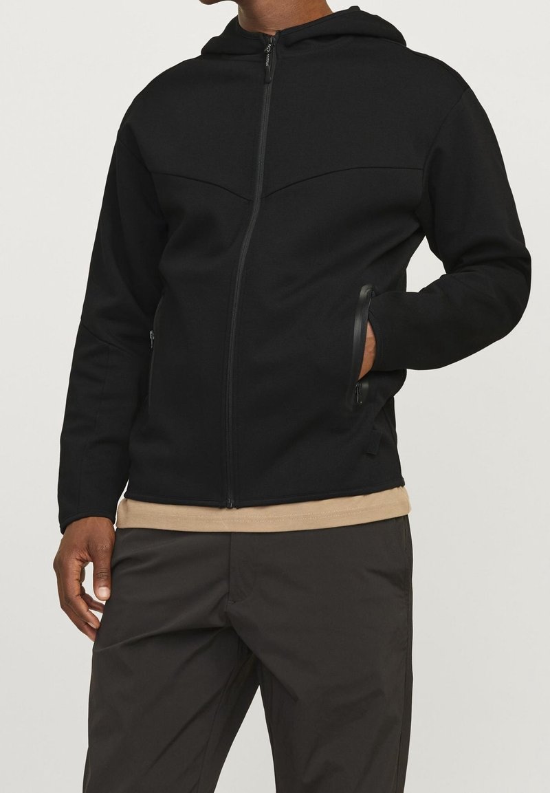 Homme portant une veste à capuche noire zippée, main dans la poche latérale, chemise beige en dessous et pantalon foncé sur fond neutre.