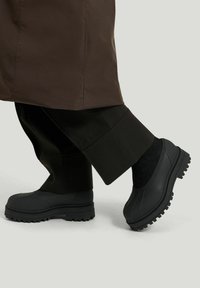 Person som bär svarta klumpiga gummistövlar med tjocka sulor och mörka vida byxor mot en neutral bakgrund.