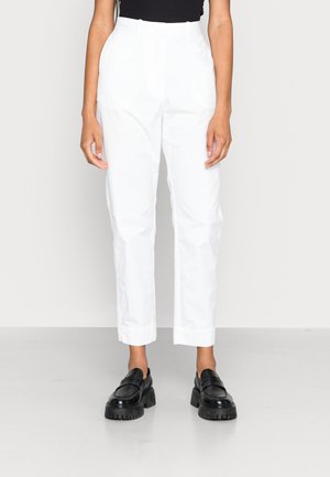 Pantalon en coton blanc avec une coupe droite et taille haute. Porté avec des mocassins noirs épais sur un fond uni.
