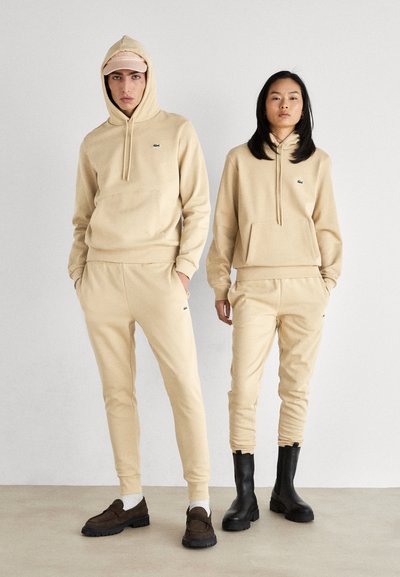 Sweatshirts à capuche beiges avec poches avant, assortis à des joggings. Des chaussures foncées ajoutent un contraste. Logo Lacoste visible.