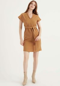 Braunes, kurzärmeliges Kleid mit V-Ausschnitt, tailliert und mit passendem Gürtel. Glattes Material mit eleganter Silhouette. Kombiniert mit beigen Knöchelstiefeln.