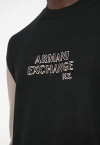 Μαύρο πλεκτό πουλόβερ με μπεζ λεπτομέρειες. Διαθέτει λογότυπο «ARMANI EXCHANGE N.Y.» με γκρι κέντημα στο εμπρόσθιο μέρος. Ριμπ λαιμόκοψη και άκρα μανικιών.