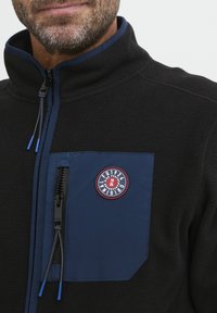 Chaqueta de forro polar negra con un bolsillo en el pecho de color azul marino, que presenta un parche de logotipo redondo y un cierre frontal con cremallera y tiradores azules.