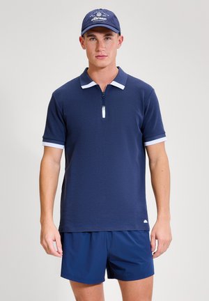 Mladý muž v tmavě modrém polo tričku s bílým lemováním a zipem, v tmavě modrých šortkách a modré čepici Ellesse, stojící před světlým pozadím.