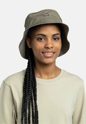 Smiling woman with long braided hair wearing a light beige bucket hat and matching crewneck shirt, facing forward against a plain background.