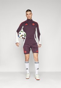 Completo da allenamento per il calcio di colore viola con maniche lunghe, dettagli rossi e logo. Include pantaloni corti e calze bianche con strisce nere. In piedi con un pallone da calcio.