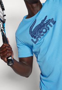Person holding tennis racket wearing bright blue Lacoste t-shirt with large crocodile logo across the chest.