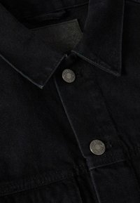 Giacca in denim nero con colletto classico, bottoni a pressione in argento e una targhetta in pelle sul colletto interno. Tessuto morbido e testurizzato.