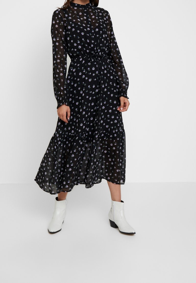 Femme portant une robe midi noire avec de petites impressions florales violettes, des manches longues transparentes et des bottines blanches, debout sur un fond blanc uni.