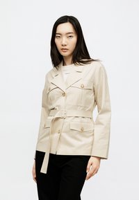 Γυναίκα με μακριά σκούρα μαλλιά, που φοράει ένα μπεζ σακάκι με ζώνη πάνω από λευκό τοπ και μαύρο παντελόνι, στέκεται μπροστά σε ένα απλό λευκό φόντο.